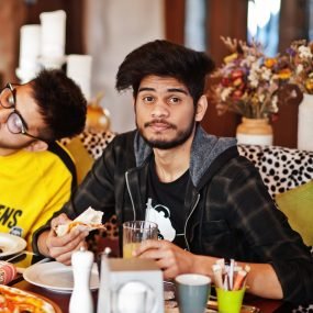 Asian friends guys eating pizza during party at pizzeria. Happy indian people having fun together, eating italian food and sitting on couch.